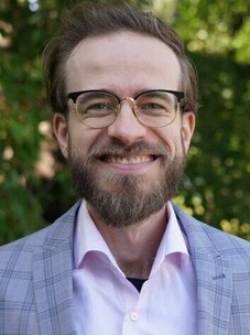 Herr Smejkal mit Brille und Bart, lächelnd, draußen vor grünem Hintergrund.